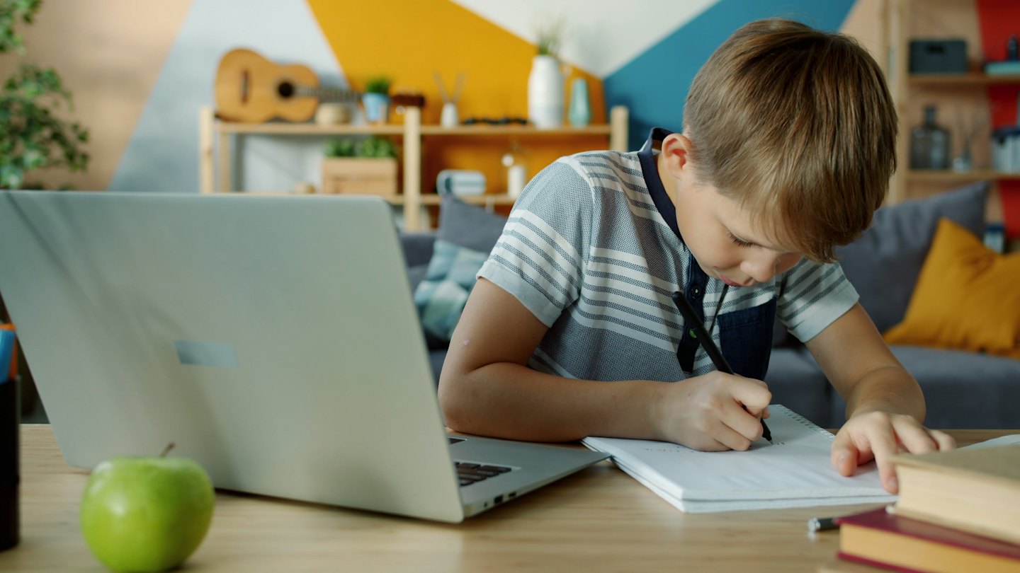 Chlapec píše v sešitu u stolu s notebookem.